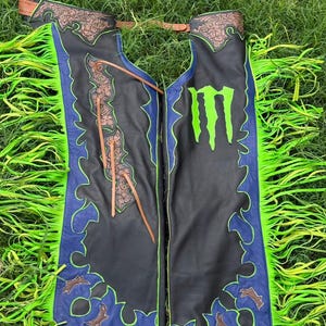 May include: Black leather chaps featuring bright neon green fringe along the outer edges, blue leather trim, and a large neon green Monster Energy logo on the right leg. The upper sections include brown floral-tooled leather accents and leather ties.