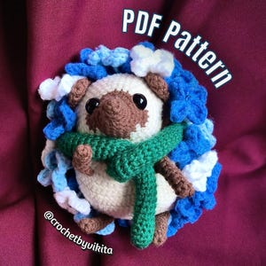 May include: A crocheted hedgehog with a cream-colored body, brown snout, and black eyes. It has a green scarf and is surrounded by blue and white crocheted accents. The text "PDF Pattern" and "@crochetbyvikita" are visible.