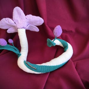 Op de afbeelding: Een handgemaakte gehaakte bloem in tinten paars, wit en blauwgroen. De grote bloemkop is paars, met een witte stengel en een blauwgroen en wit gestreepte rank. Het ontwerp bevat twee paarse knoppen en groene bladeren. Het item is een decoratief stuk.