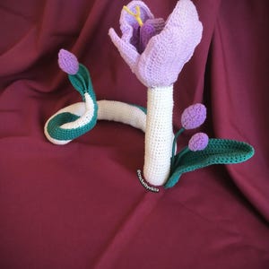 Op de afbeelding: Gehaakte bloem- en slangsculptuur. De bloem is lavendelkleurig met een witte stengel en groene bladeren. De slang is wit met groen-witte strepen. De sculptuur staat tegen een bordeauxrode achtergrond.