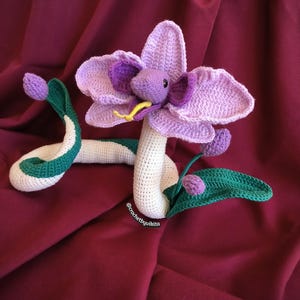 Op de afbeelding: Een gehaakte slang met een bloemhoofd. De slang is wit met groene en witte accenten. De bloem is paars en lavendelkleurig met een gele tong. De achtergrond is bordeauxrood. De tekst "@crochetbyvikita" is zichtbaar.