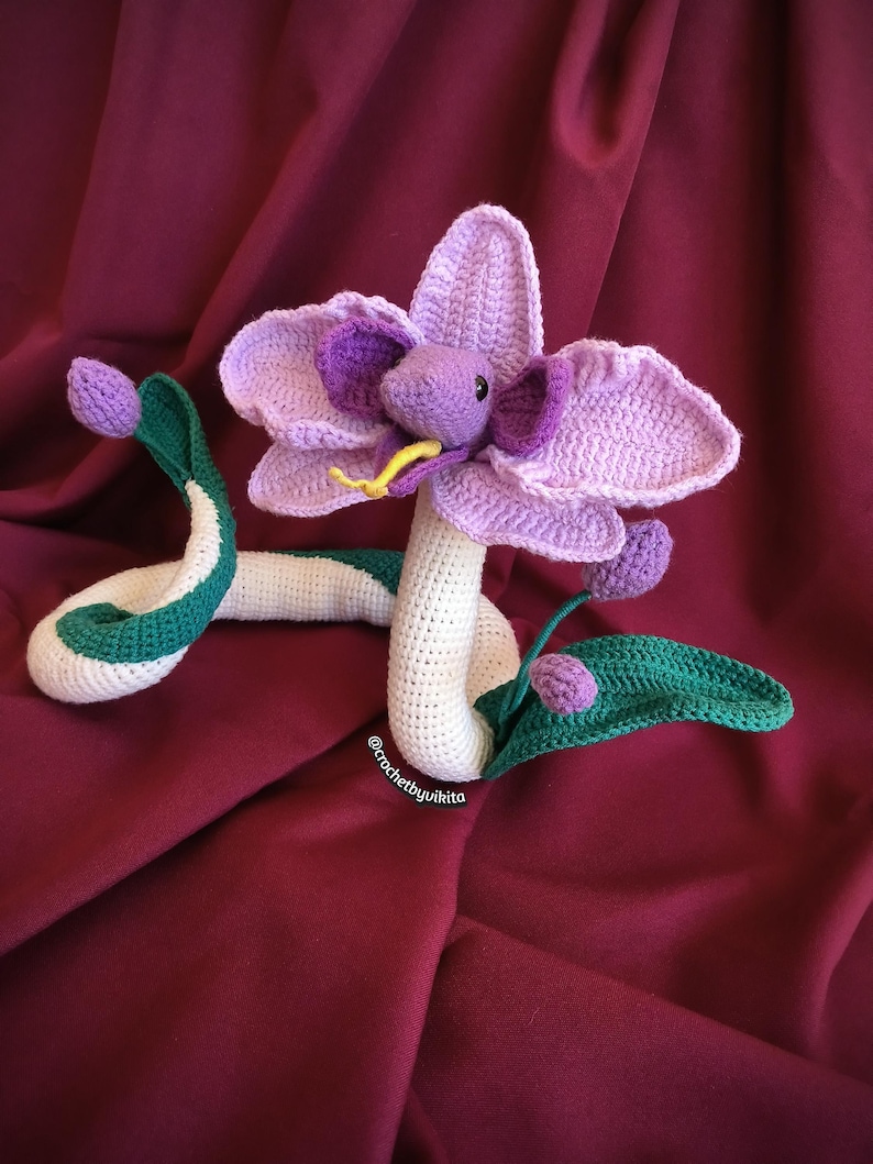 Op de afbeelding: Een gehaakte slang met een bloemhoofd. De slang is wit met groene en witte accenten. De bloem is paars en lavendelkleurig met een gele tong. De achtergrond is bordeauxrood. De tekst "@crochetbyvikita" is zichtbaar.