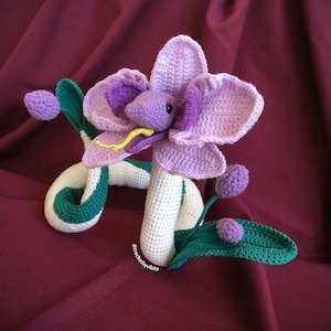 Op de afbeelding: Handgemaakte gehaakte bloemenslang in paars, groen en wit. De bloem heeft grote bloemblaadjes en een slangachtig lichaam met groene accenten. De tong van de slang is geel. Een uniek decoratief item.