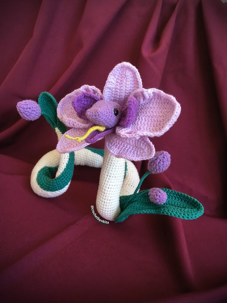 Op de afbeelding: Handgemaakte gehaakte bloemenslang in paars, groen en wit. De bloem heeft grote bloemblaadjes en een slangachtig lichaam met groene accenten. De tong van de slang is geel. Een uniek decoratief item.