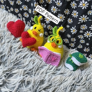 Puede incluir: Colección de artículos de ganchillo hechos a mano, que incluyen figuras amarillas con forma de pollito con detalles verdes, corazones rojos y un pequeño objeto verde y blanco. El texto "PDF Pattern" es visible. Los artículos están sobre una superficie blanca y gris esponjosa.