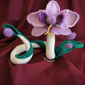Op de afbeelding: Een gehaakte sculptuur van een bloem-slang hybride. De bloem heeft lavendelkleurige bloemblaadjes en een paars centrum, met een gele tong. Het slangenlichaam is wit met groene strepen. De sculptuur staat op een bordeauxrode achtergrond. De tekst "@crochetbyvikita" is zichtbaar.