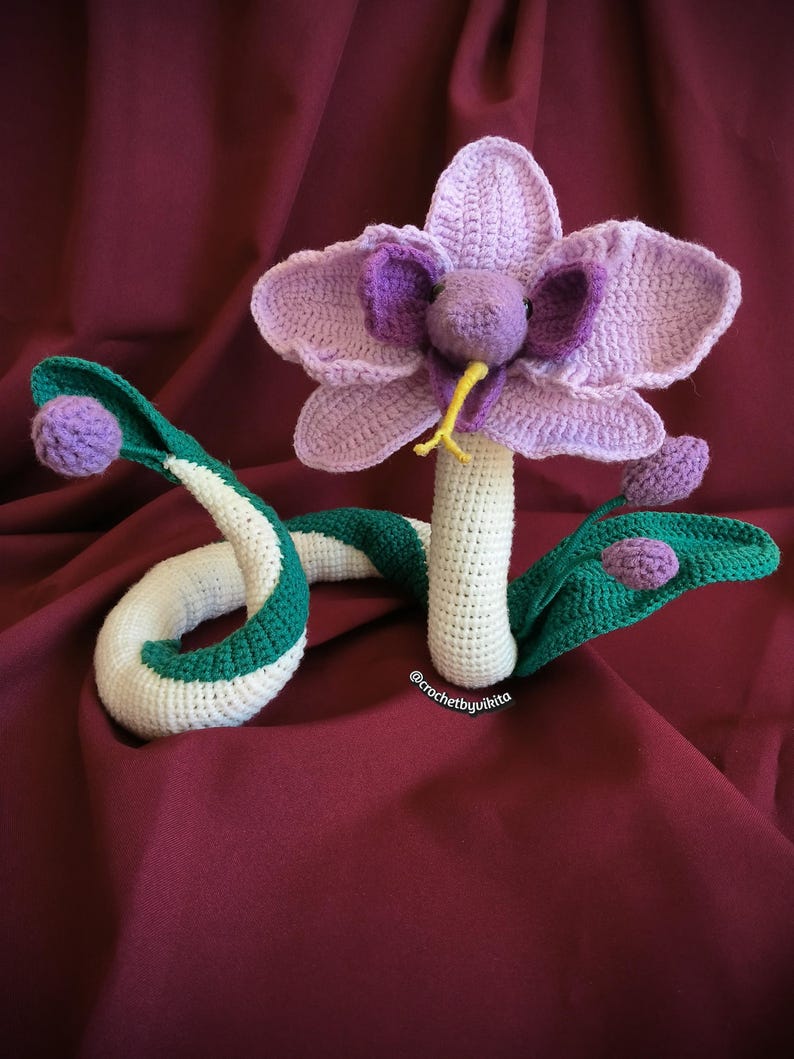 Op de afbeelding: Een gehaakte sculptuur van een bloem-slang hybride. De bloem heeft lavendelkleurige bloemblaadjes en een paars centrum, met een gele tong. Het slangenlichaam is wit met groene strepen. De sculptuur staat op een bordeauxrode achtergrond. De tekst "@crochetbyvikita" is zichtbaar.