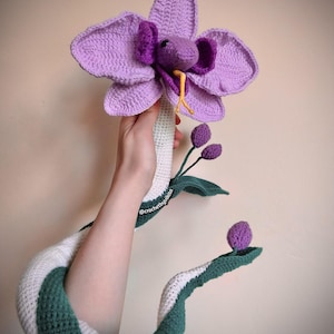 Op de afbeelding: Een handgemaakte gehaakte orchidee met een slangenontwerp. De bloem is levendig paars, met een slangenkop die uit het midden komt. Het lichaam van de slang is wit en groen, en de stengel heeft groene bladeren en paarse knoppen.