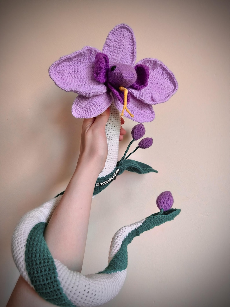 Op de afbeelding: Een handgemaakte gehaakte orchidee met een slangenontwerp. De bloem is levendig paars, met een slangenkop die uit het midden komt. Het lichaam van de slang is wit en groen, en de stengel heeft groene bladeren en paarse knoppen.
