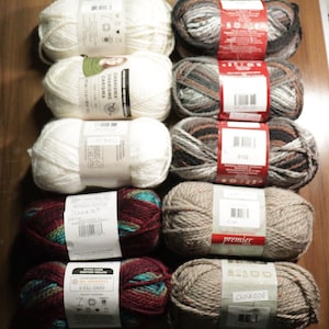 May include: An assortment of yarn skeins in various colors. The selection includes white, gray, brown, and multicolored skeins. Some skeins have labels with the brand name and product information. The yarn is arranged on a wooden surface.