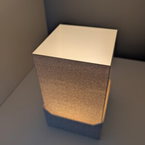 May include: A small, square lamp with a textured, metallic-looking shade. The top of the lamp is a solid, off-white color. The lamp is sitting on a white surface, and the background is a neutral color.