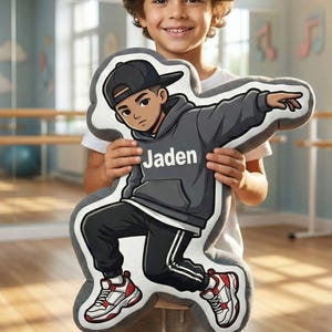 Może przedstawiać: Pluszowa poduszka w kształcie kreskówkowego tancerza, w szarej bluzie z kapturem i czarnych spodniach, z imieniem "Jaden" z przodu. Tancerz ma na sobie czapkę z daszkiem i czerwono-białe trampki. Poduszkę trzyma uśmiechnięte dziecko.