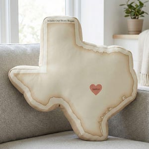 Puede incluir: Un cojín decorativo con forma del estado de Texas, con un diseño de mapa vintage. El cojín tiene un corazón con la palabra "Austin" y el texto "Where Our Story Began". Beige con bordes marrones.