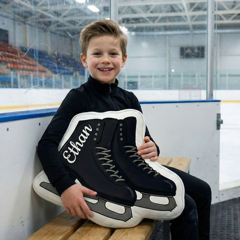 May include: A decorative pillow in the shape of ice skates. The pillow is black with white accents and gray blades. The name "Ethan" is printed in white script on the side of one skate. The pillow is being held by a person.