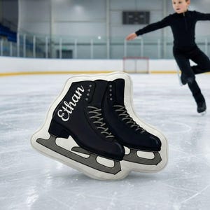 May include: A decorative pillow shaped like a pair of black ice skates with silver blades. The name "Ethan" is printed in white script on the side of one skate. The pillow is on an ice rink.