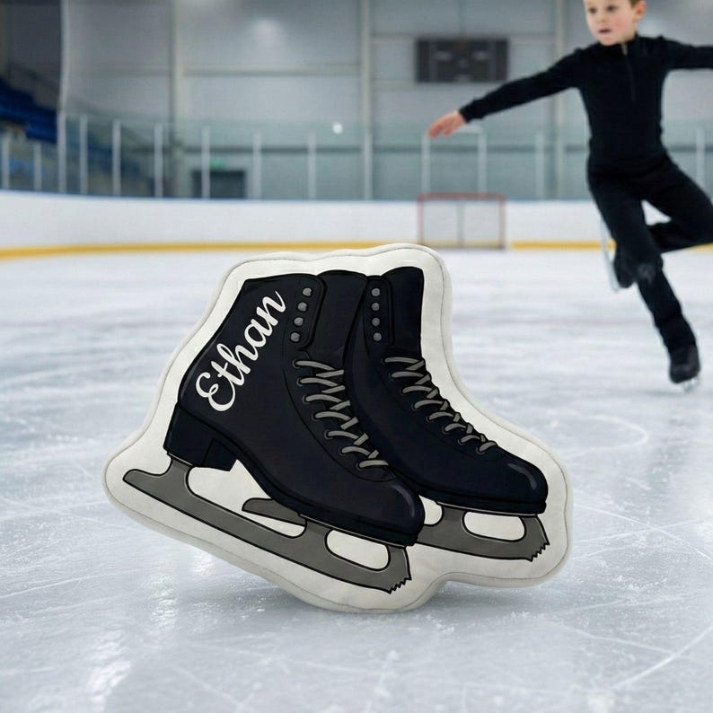 May include: A decorative pillow shaped like a pair of black ice skates with silver blades. The name "Ethan" is printed in white script on the side of one skate. The pillow is on an ice rink.