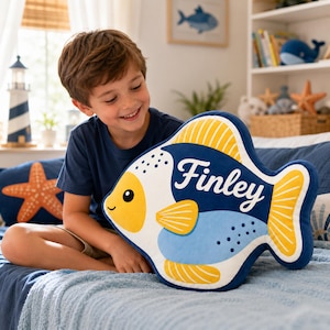 May include: A plush fish-shaped pillow featuring a dark blue, light blue, white, and yellow color scheme. The pillow displays the name Finley in white script lettering on its side, with yellow fins and tail, and a white face with a black eye.