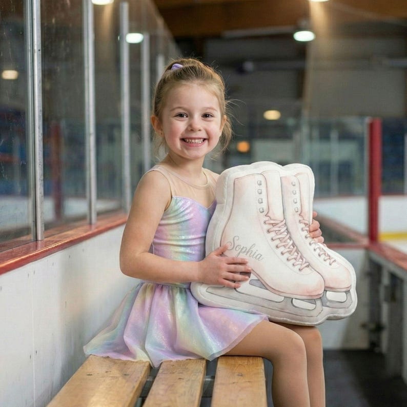 May include: A young girl smiles while holding a decorative ice skate pillow. The pillow is white with pink laces and the name "Sophia" printed on the side. The girl is wearing a pastel rainbow dress.