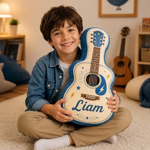 Könnte beinhalten: Ein gitarrenförmiges Plüschkissen in Creme und Blau mit aufgedrucktem Griffbrett, Schallloch und Blitzmotiven. Der Name Liam ist in einer dunkelblauen Schreibschrift auf dem unteren Teil des gitarrenförmigen Kissens aufgedruckt.