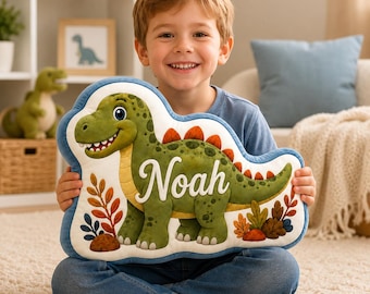 May include: A plush, die-cut cushion shaped like a cartoon dinosaur with green skin, orange spikes, and a cream-coloured belly. The dinosaur is printed on a white background with colourful foliage and the name Noah written in white script across its body.