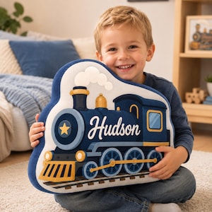 May include: A plush pillow shaped like a steam locomotive train. The train is dark blue with gold accents, a yellow star on the front, and the name Hudson printed in white script. It features a white smoke cloud and sits on train tracks.