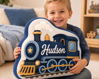 May include: A plush pillow shaped like a steam locomotive train. The train is dark blue with gold accents, a yellow star on the front, and the name Hudson printed in white script. It features a white smoke cloud and sits on train tracks.