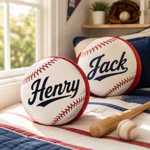 Puede incluir: Dos cojines redondos de peluche con temática de béisbol, blancos con detalles de costuras rojas. Un cojín lleva el nombre Henry en letra cursiva azul oscuro, y el otro lleva el nombre Jack en letra cursiva azul oscuro. Están sobre una cama con un bate.