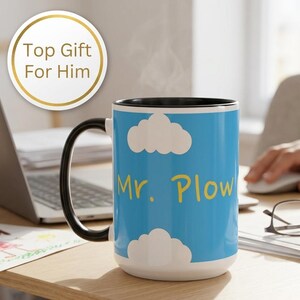 Op de afbeelding: Een keramische mok met een zwarte handgreep en binnenkant, voorzien van een lichtblauwe buitenkant met witte wolken en de gele tekst Mr. Plow in het midden gedrukt. De mok staat op een houten bureau bij een laptop en een bril.