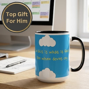 May include: A ceramic mug with a black handle and interior, featuring a bright blue exterior with white clouds and the yellow text "So this is what it feels like when doves cry" printed on the front. The mug has a cylindrical shape.