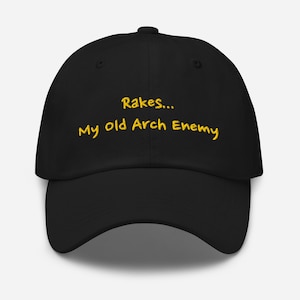 May include: Black baseball cap with the text "Rakes... My old Arch Enemy" in yellow script. The cap has a curved brim and a button on top. A casual accessory for outdoor activities or everyday wear.