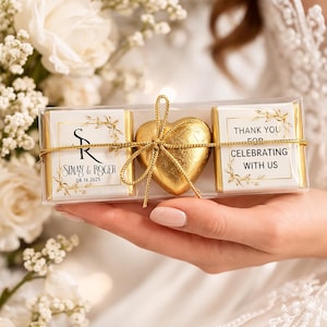 May include: A clear box containing a gold heart-shaped chocolate and two rectangular chocolates. The rectangular chocolates have gold floral designs and text that reads "SIMAN & ROGER 08.18.2025" and "THANK YOU FOR CELEBRATING WITH US". The box is tied with gold string.