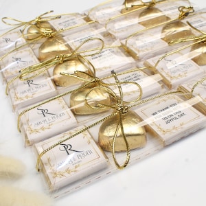 May include: Favours wrapped in clear plastic with gold detailing. Each contains a gold-wrapped chocolate and a card with the text "SARAH & ROGER" and "WE THANK YOU FOR BEING WITH US ON THIS JOYFUL DAY."