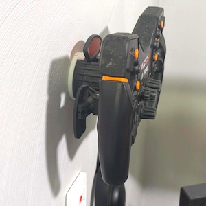 May include: A black racing steering wheel controller mounted to a wall, featuring orange buttons, rotary dials, and paddle shifters. The device has a textured black finish and is secured by a circular white mounting bracket attached to the wall surface.