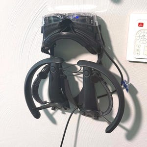 May include: A black virtual reality headset with a clear front panel and two matching black handheld controllers with circular tracking rings, all mounted together on a wall-mounted storage bracket, positioned next to a white wall light switch.
