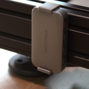 May include: A silver Insignia USB 3.0 hub with a rectangular body and rounded corners, featuring the brand name printed vertically on the front. The device is secured to a black metal extrusion frame by two grey 3D-printed mounting clips.