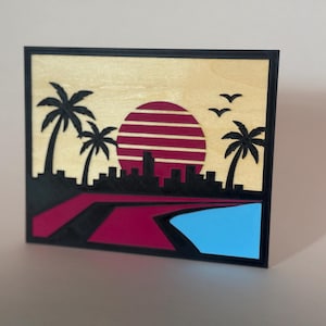 Puede incluir: Un cuadro rectangular de madera con un diseño en capas de una puesta de sol sobre el horizonte de una ciudad. La escena incluye siluetas negras de palmeras, pájaros y edificios sobre un fondo de madera clara con un sol rojo a rayas.