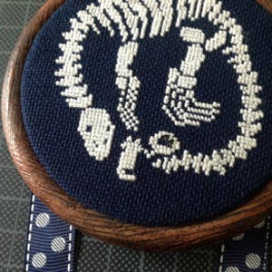 以下が含まれることがあります： 濃い青色の円形背景に、白い刺繍で描かれたロブスターの骨格。木製のフレーム付き。