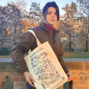 Può includere: Una borsa tote beige con la scritta "PALEONTOLOGY" stampata in alto. La borsa presenta illustrazioni nere di fossili di dinosauri. La borsa è tenuta davanti a un muro di mattoni e alberi.