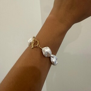 May include: A gold bracelet with large, irregularly shaped white pearls. The bracelet has a toggle clasp and is worn on an arm. The pearls have a lustrous sheen, and the bracelet is set against a neutral background.