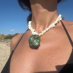May include: A necklace featuring a large, green and brown sea shell pendant. The pendant is suspended from a necklace made of small, white shell fragments. The background is a sunny beach scene with a blue sky.
