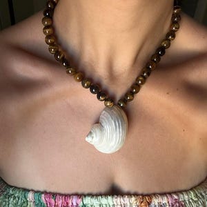 Op de afbeelding: Een ketting bestaande uit een reeks ronde, gepolijste bruine en gouden tijgeroogkralen, gecentreerd met een enkele, gladde, gebroken witte spiraalvormige schelphanger die aan de basis van de nek bij het sleutelbeen hangt.