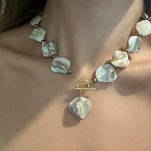 Può includere: Collana con grandi perle di conchiglia irregolari nei toni crema, grigio e beige. La collana ha una chiusura e un ciondolo color oro. Le perle sono infilate con piccole perle color oro.