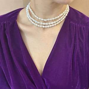 May include: A triple-strand pearl necklace is worn over a deep purple velvet top. The necklace features small, off-white pearls. The top has a V-neck design and long sleeves. The image is a close-up of the neck and upper chest.