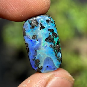 May include: A polished opal gemstone with a rectangular shape. The opal displays a vibrant play of color, with blues, greens, and hints of white. Black matrix patterns are scattered across the surface, adding contrast and depth to the stone.