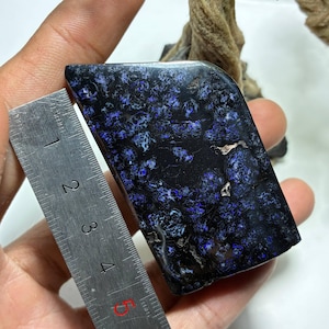 May include: A rectangular gemstone with a polished surface, featuring a dark base color with intricate patterns of blue and white. A ruler is placed next to the stone for measurement. The stone's dimensions are approximately 4 inches in length.