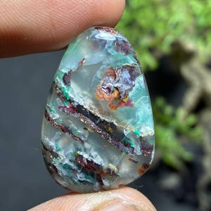 May include: A teardrop-shaped gemstone with a translucent, light blue base. The stone features intricate patterns of dark brown, red, green, and purple inclusions. The gemstone is held between two fingers, showcasing its polished surface and natural beauty.