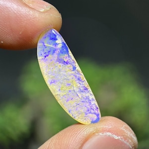 May include: A polished, elongated gemstone with a blend of colors. The stone features a base of pale yellow, overlaid with patches of lavender and streaks of deep blue. The gemstone is held between two fingers.