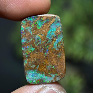 May include: A rectangular boulder opal gemstone featuring a natural brown ironstone matrix with vibrant flashes of iridescent green and blue color play distributed across the surface in a mottled, organic pattern with a polished finish.