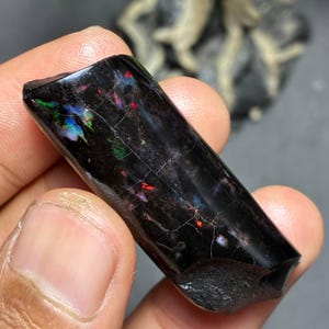 May include: A polished black opal gemstone with vibrant flashes of color, including green, blue, and red. The rectangular stone has a glossy surface with visible internal fractures, showcasing its natural beauty. The opal is a unique piece of jewelry.