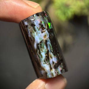 May include: A polished, rectangular black opal gemstone with iridescent flashes of green, blue, and red. The opal's surface has a unique pattern of light blue and white swirls, creating a striking visual effect. The gemstone is held between fingers.
